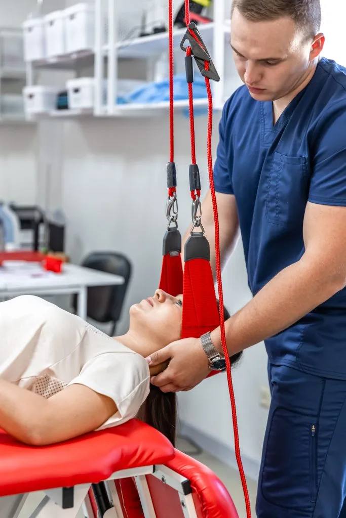 Physical therapist in blue scrubs adjusting suspension straps for patient lying on red treatment table