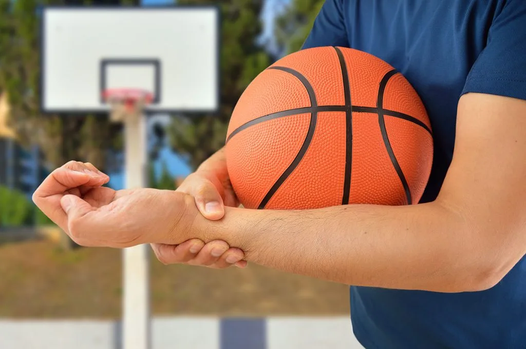 Basketball player in blue shirt holding orange ball, cradling wrist with opposite hand in pain near outdoor court