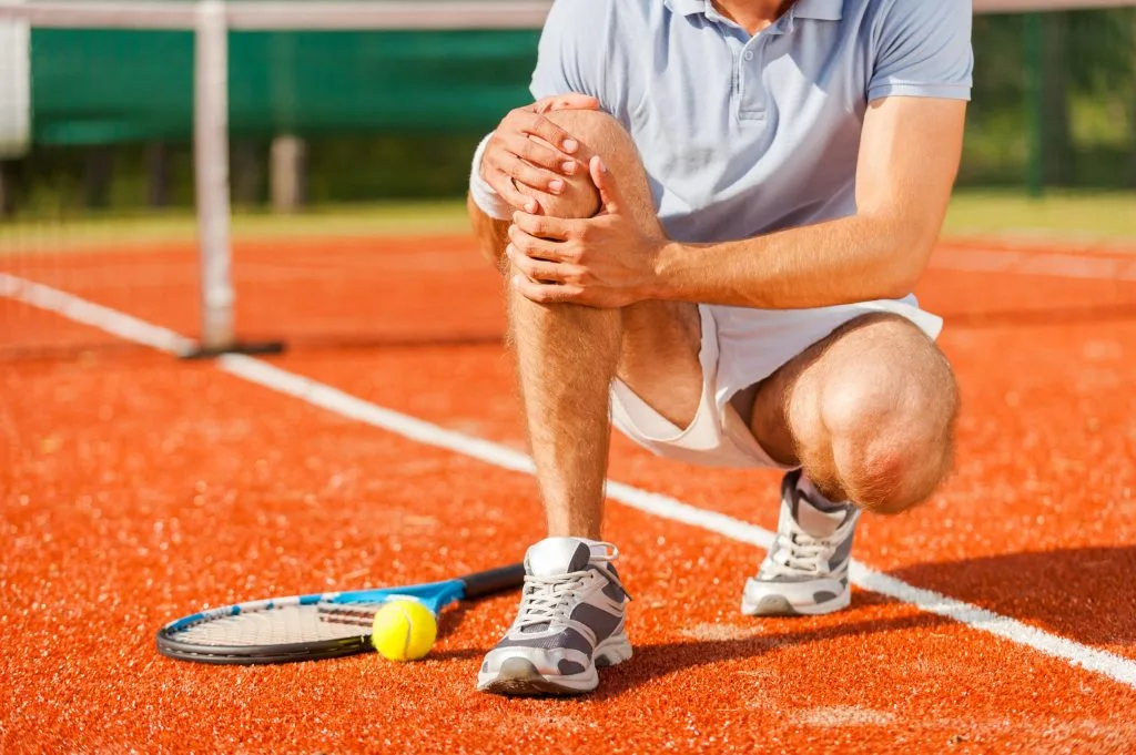Tennis player crouching on orange clay court, holding knee with both hands while racket and ball lie nearby