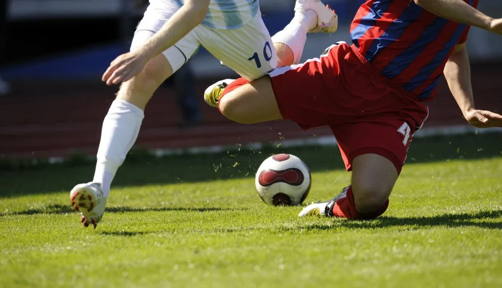 Two soccer players competing for ball on grass field, one in white and one in red striped uniform
