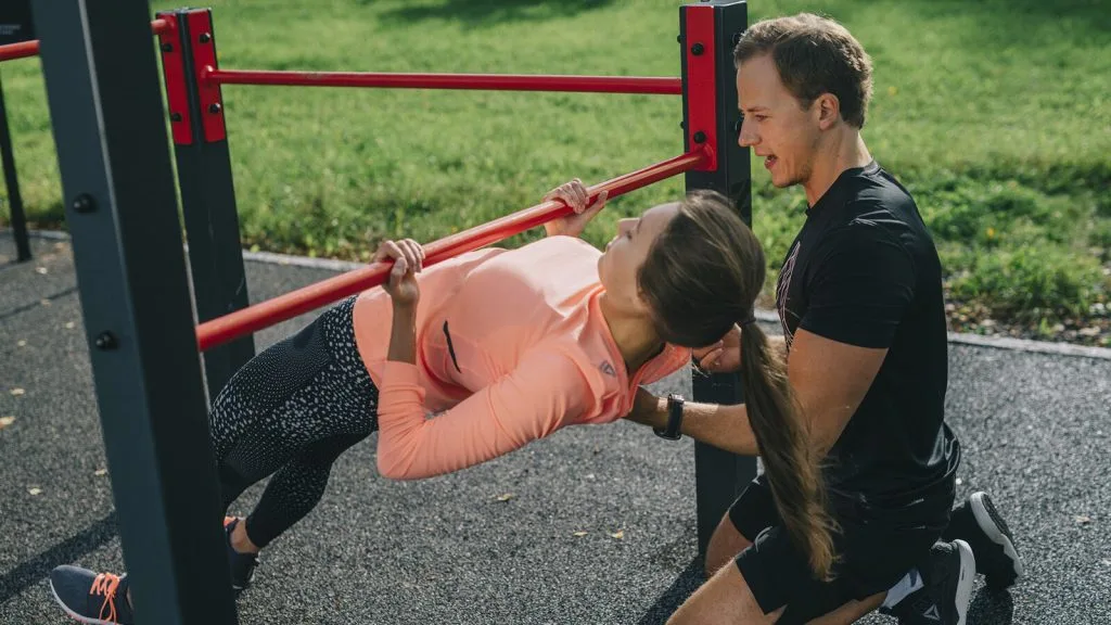 Female trainer assisting woman performing push-up against red horizontal bar in outdoor fitness setting