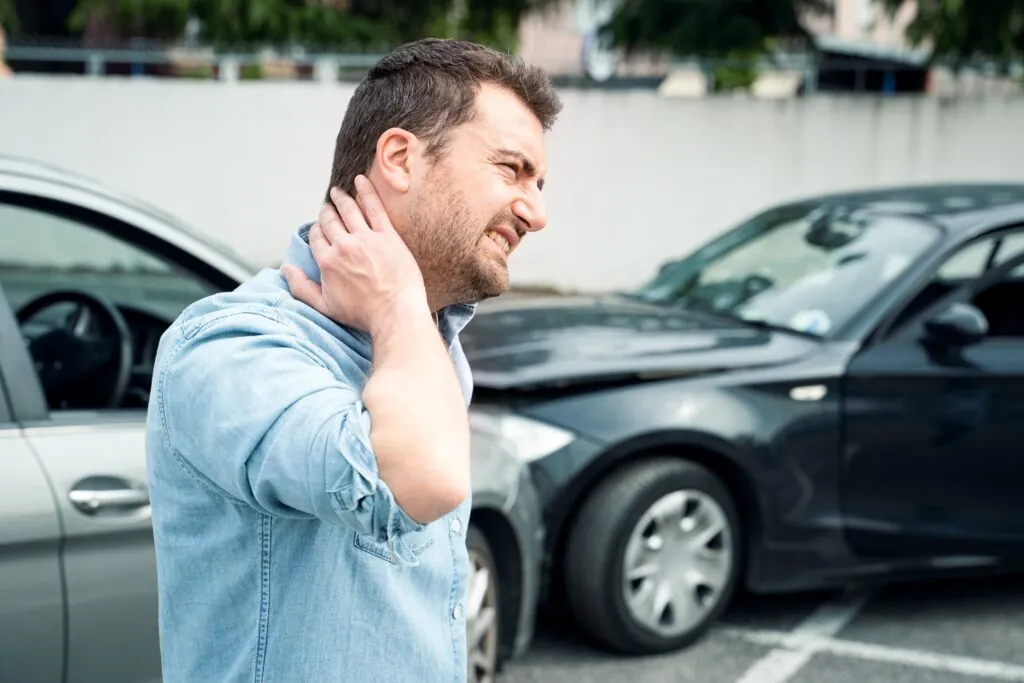 Man touching neck in pain at parking lot with damaged cars visible