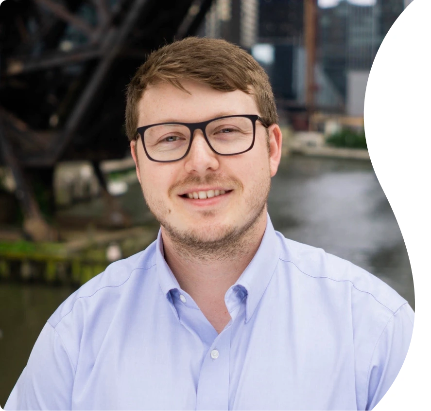 Man wearing glasses and light blue button-up shirt, smiling at camera with urban waterfront in background