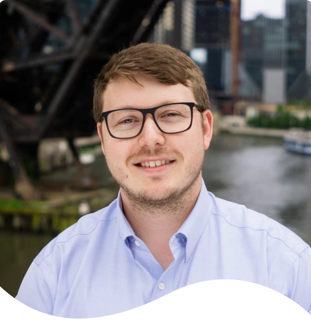 Man wearing glasses and light blue shirt smiling at camera with urban waterfront in background