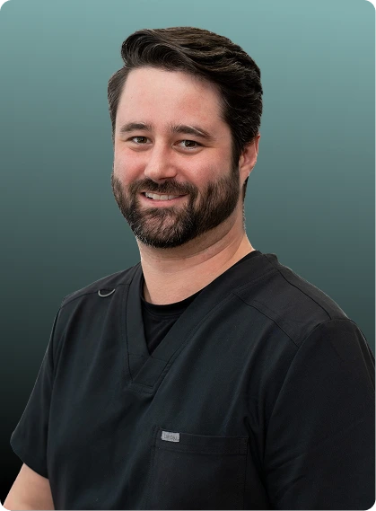 Man with dark hair and beard wearing black scrubs, smiling at camera against teal background