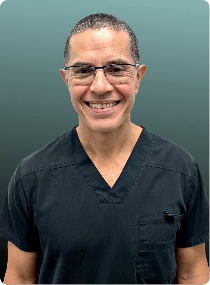 Man wearing glasses and black v-neck scrubs, smiling at camera against teal background
