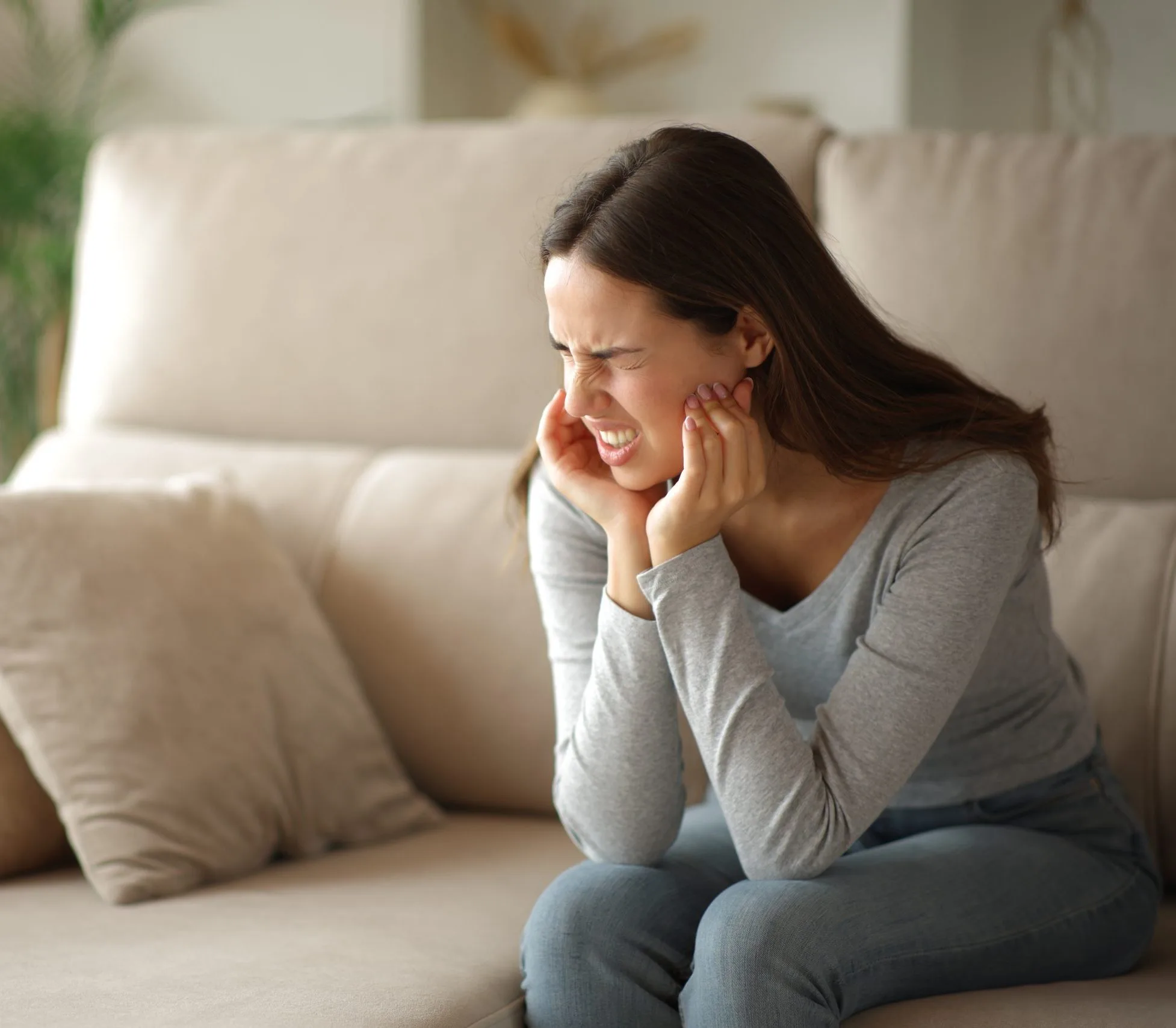 Woman sitting on a couch holding her jaw in pain, illustrating TMJ discomfort